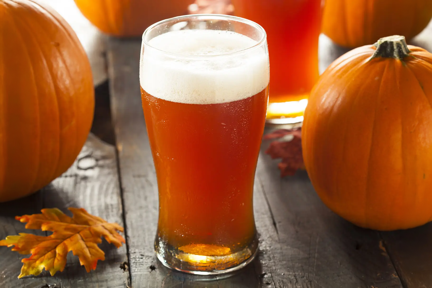 A pint of beer on a wood table surrounded by pumpkins and a leaf.